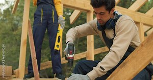 man contractor in work clothes sit on beam install new roofing tools, using electric drill building cottage house Building Technologies. builder with building level standing in background