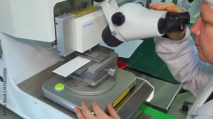 Quality control employee inspects a manufactured LED panel for defects using microscope. Research lab scientist examines an LED board through a microscope. Researcher in white coat at his workstation.