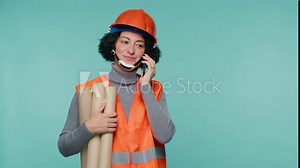 Mature female engineer in uniform and hardhat standing technical blueprint rolls at teal blue studio background. Industrial construction architect specialist talking with client using smartphone 4K
