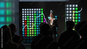 Woman presenting project on stage during meeting. Shot from audience, people listening businesswoman speaker with tablet talks with audience answers questions