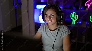 Confident young female gamer, a radiant blonde streamer, cheerfully flashes a heart symbol with her hands during a engaging night stream in her home-based gaming room