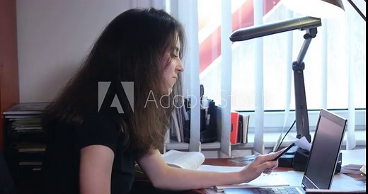 A close shot of cute and focused young girl using a laptop computer and writing on notebook, learning and doing homework sitting on the platform