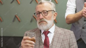 Professional young barber giving a haircut with concentration to a rich respectable gentleman, standing and holding razor while relaxed elderly client is smoking cigar and drinking alcohol.