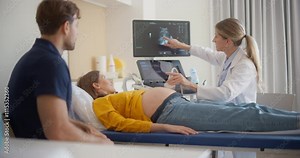 Caring Female Hospital Obstetrician Conducting an Ultrasound Exam for a Pregnant Woman. Young Couple Watching the Screen while Doctor Describes the Baby’s Growth to Expectant Mother