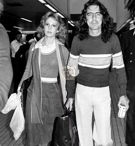"Shock rocker Alice Cooper and his future wife, Sheryl Goddard, were captured in a rare candid moment at London’s Heathrow Airport in 1975. At the time, Cooper was promoting his solo album Welcome to My Nightmare, while Sheryl performed as a dancer in the stage production that accompanied his theatrical tour. The two had met just a year prior, and their bond quickly deepened behind the scenes. Within months of this photo—circa March 1976—they were married in a quiet ceremony, marking the start o