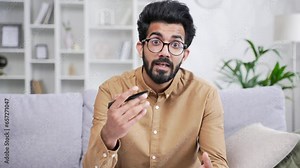 Webcam view. Confident teacher in glasses talking on video call while sitting in home office looking at camera. A young coach or tutor conducts an online training, seminar, course or webinar remotely