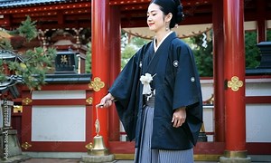 A Shinto priest in traditional robes stands before a sacred shrine gate (torii) holding a ritual object. The shrine is red and white with trees and a water stream in the background.