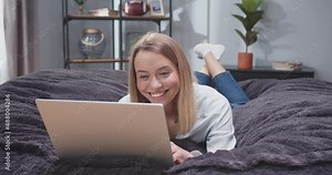 Close up front view portrait of adult woman lying on her stomach on sofa with laptop. Female scrolling social media on gadget and laughing. Woman after work resting on sofa with computer.