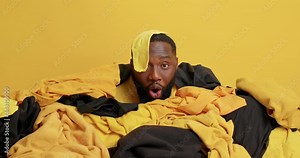 Shocked black bearded man drowned in pile of clothes collects laundry for washing does everyday routine stunned to notice something amazed isolated over yellow background. Housework and cleaning Stock Video
