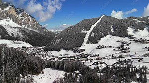 Alta Badia, Italy: Aerial view of ski resort in snowy Dolomites. Charming town landscape with chalets and hotels surrounded by snow-capped peaks, sunny day, blue sky