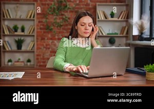 A woman in a green sweater sits at a desk, appearing stressed or tired, in a modern office setting with a laptop and documents Stock Video Footage - Alamy