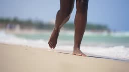 Black young fit woman's legs walking barefoot along a sunny beach,...