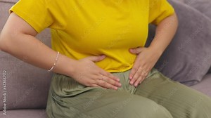 Stomach pain, hands and woman on a sofa with pms, cramps or gas, menstruation or bloating in her home. Tummy, ache and female person with endometriosis, fibroids and colon or constipation problem