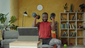 Fit strong muscular man lifting up heavy dumbbells, working out biceps exercises, watching online training lessons on laptop. Home isolation. Bodybuilding.