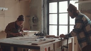 Lockdown of young female carpenter and her male coworker standing at workbench in joinery workshop and carving wood pieces with cutters