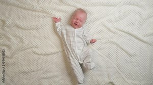Sleepy sad newborn baby is crying in bed. Upset baby boy shouting and grimacing face - bedtime problems parenting.