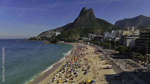 Backward aerial over crowded and bustlin Lebron Beach in Rio de Janeiro, Brazil, overlooking the iconic Pedra do Arpoador