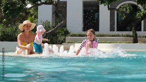 Mother with kids having fun in swimming pool, slow motion. Young woman holding cute baby in hands while older daughter splashing in water