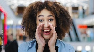Mysterious african american woman with curly hair keeps index finger on lips tells secret makes silence gesture quiet sign talking secrecy information whispers gossip mystery indoor looking at camera