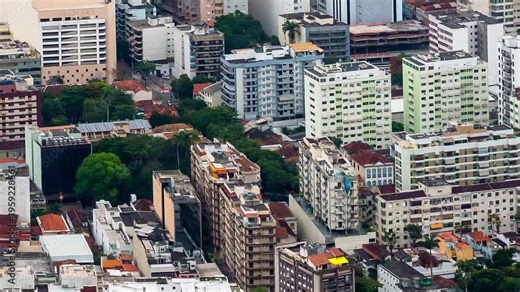 Time-lapse footage of a densely populated urban area captured from above. Numerous mid-rise residential and commercial buildings of varying architectural styles and colors fill the compact city layout
