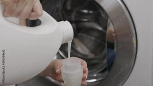 Against background of drum of steel-colored washing machine, woman pours liquid washing gel into plastic cap. A girl in a white T-shirt carefully pours a transparent conditioner for flattening laundry