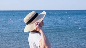 Happy mature woman 50 years old dressed in white dress, straw beach and sunglasses on the beach near the seashore. Vacation concept, relaxation, retirement age