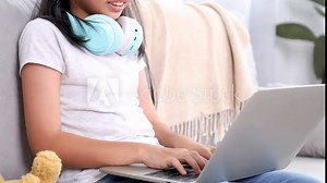 Young Asian elementary girl doing homework from school at home on weekend, schoolkids student happy using technology laptop computer learning or online meeting with friends for assignment group report