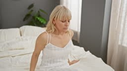 A contemplative blonde woman in a white nightgown sits on a bed in a...
