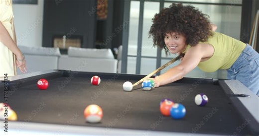 African American woman playing pool with friend at home, taking turn lining up cue to score