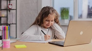 Cute primary school girl studying at home using laptop computer. Schoolgirl has online lesson, writing in exercise book, doing homework. Distance learning online. Children home education