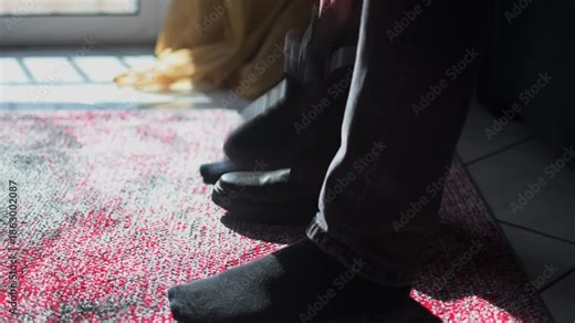Close-up static shot footage shows a person wearing gray jeans tying his shoes in a room with a carpet. Light of enters the room from the window.