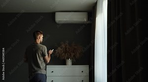 Man tries to turn on air conditioner using remote control in apartment, static shot. Slender guy presses buttons, climate control does not work. Frustrated person leaves.