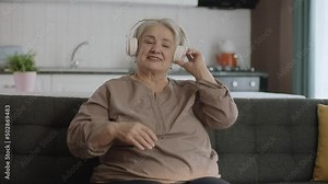 Mature cheerful woman enjoying life after retirement.Colorful and funny moments of an old woman listening to music with headphones on her sofa. Funny old woman with headphones singing and dancing.