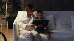 Grandmother and grandson are sitting on a white sofa and reading a book together. A cozy evening at Grandma's. Senior lady and her grandson spend time together.