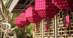 Background video of Chinese traditional red paper lanterns hanging on a wooden house with an ornament. The lanterns are swinging in the wind, with a green tree in the background. Chinese New Year.