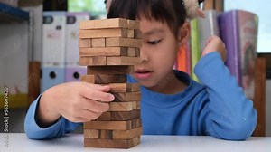 Cute little girl playing with wooden game (jenga) on a table in a room at home. Asian child playing wood blocks stack game indoors. Learning and development of fine motor skills.