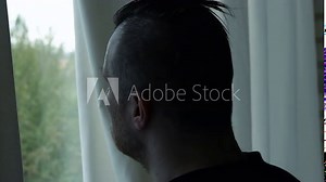 A man looks out the window. Looking out the window from behind the curtains. A young man looks at the weather outside from his apartment