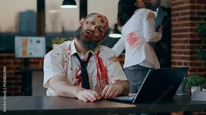Bizarre looking zombie using modern laptop in office workspace. Evil apocalyptic brain-eating monster with deep and bloody wounds and scars trying to use portable computer at work.