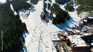 People Skiing and Riding Chairlifts down Ski Slope next to Parking Lot and Pine Tree Forest and Snow. Aerial Drone Footage