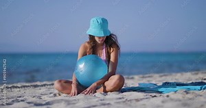 Young woman in blue hat sitting with a blue ball on a sandy beach under clear blue skies