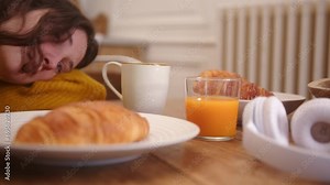 Slow dolly lateral shot of a table set for breakfast, a pair of headphones and a girl lying on the table with eyes open. She has long brown hair. Blurred background