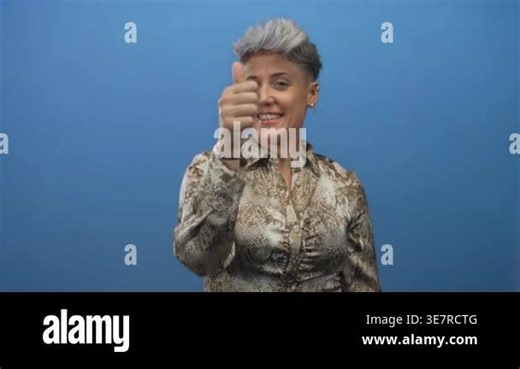 Woman with short hair wearing snake print shirt giving thumbs up gesture in studio with blue background; optimism Stock Video Footage - Alamy