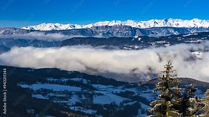 Timelapse or time lapse footage shows clouds and mountains covered with snow on their peaks. Underneath the moving clouds and between the hills and mountains there is a small town or a village in 4K.