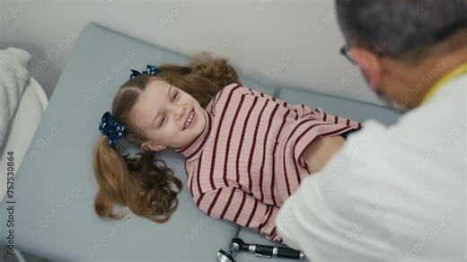Over-the-shoulder shot. A little girl patient lies on her back on the medical examination couch and laughs while her abdomen is examined by a pediatrician. A male doctor palpation a child's abdomen