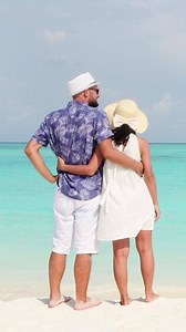 Back view candid beautiful tourist caucasian couple in white summer clothes and hats stands on a tropical beach and gazes at turquoise ocean during their vacation time honeymoon