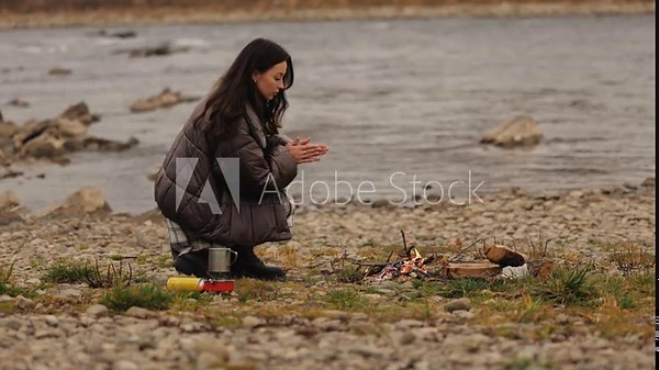 Woman building a campfire by the river during an autumn camping trip, adding branches to the fire while preparing an outdoor cooking spot in a natural setting