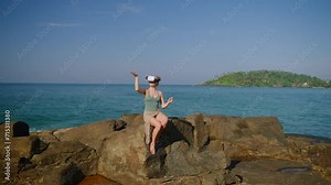 Woman in swimsuit uses VR headset on rocky seaside, experiences virtual reality with hand gestures, immersed in simulation, tech meets nature concept, interactive simulation interface in focus.