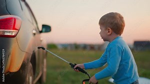 Kid washes car with sprayer in meadow. Serious little boy cleans dirty automobile with water pumps at backyard. Toddler child plays carwash in evening