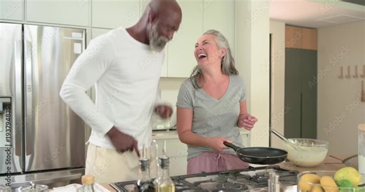 Diverse couple making pancakes at stove, woman flipping in pan using bowl while man starting dance