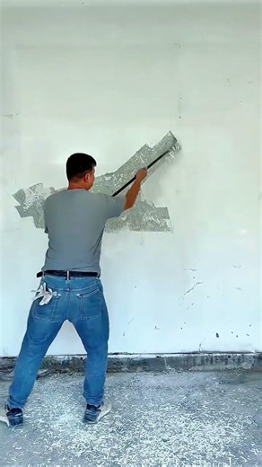 scraping a thin layer of old white paint from a large indoor wall using a long metal hand scraper